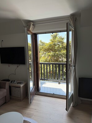 A room with a TV mounted on the wall, a sofa, a small table with remotes and a magazine, and a balcony with a railing and trees in the background.