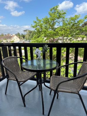Een balkon met twee rieten stoelen, een glazen tafel en een vaas met witte bloemen.
