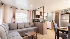 En moderne stue med sofa, sofabord og spiseplads med stole. Der er et køkken med køleskab, mikroovn og skabe.