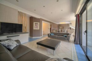 Ein modernes Wohnzimmer mit einer Couch, einem Couchtisch und einem Flachbildfernseher. Im Hintergrund ist eine Essecke mit Tisch und Stühlen zu sehen.