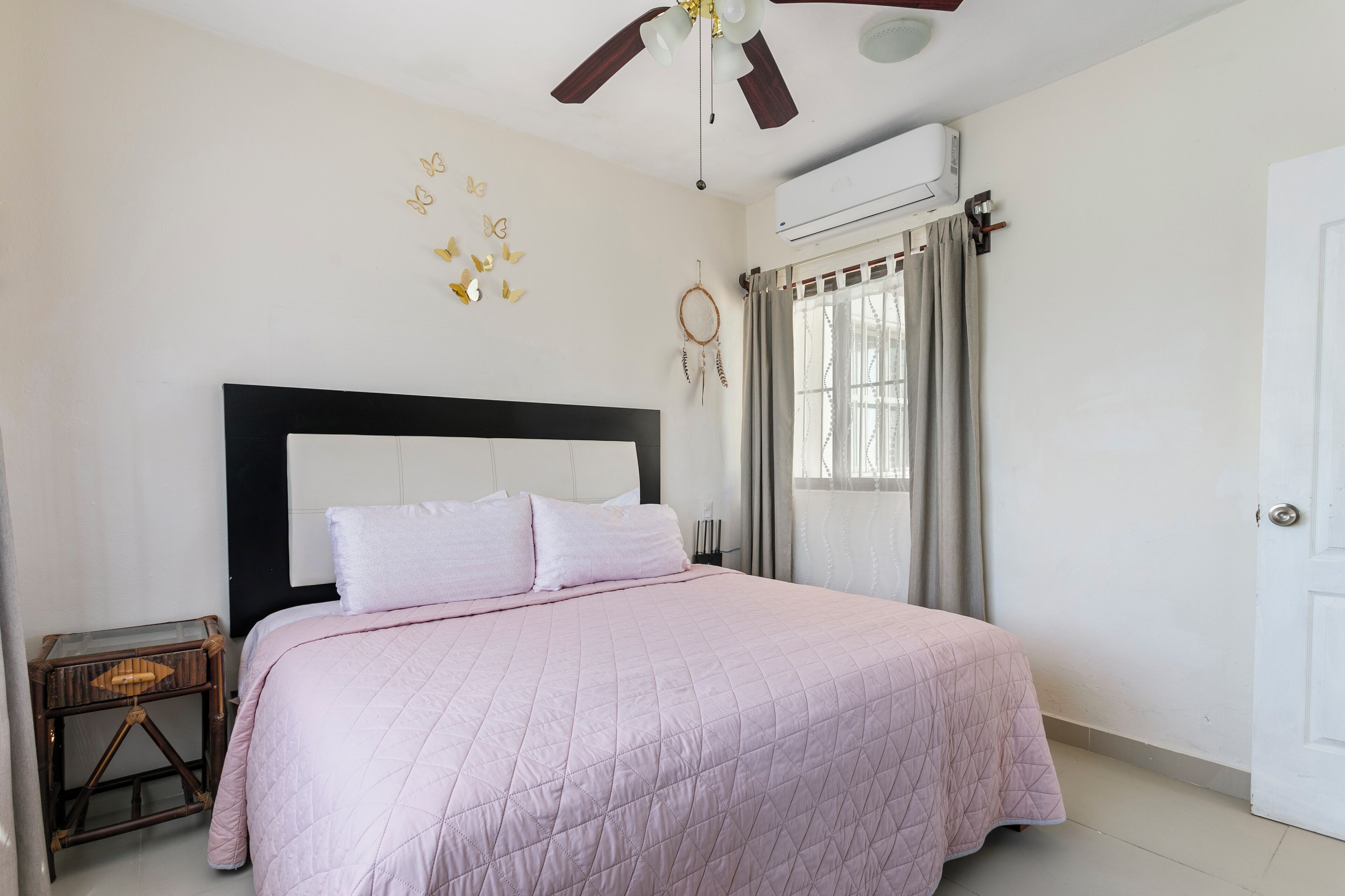 Un dormitorio con cama, ventilador de techo, aire acondicionado, mesita de noche y ventana con cortinas.