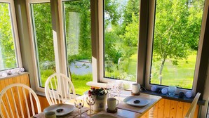 A dining area with a table set for two, white chairs, and a view of a lush garden through large windows.