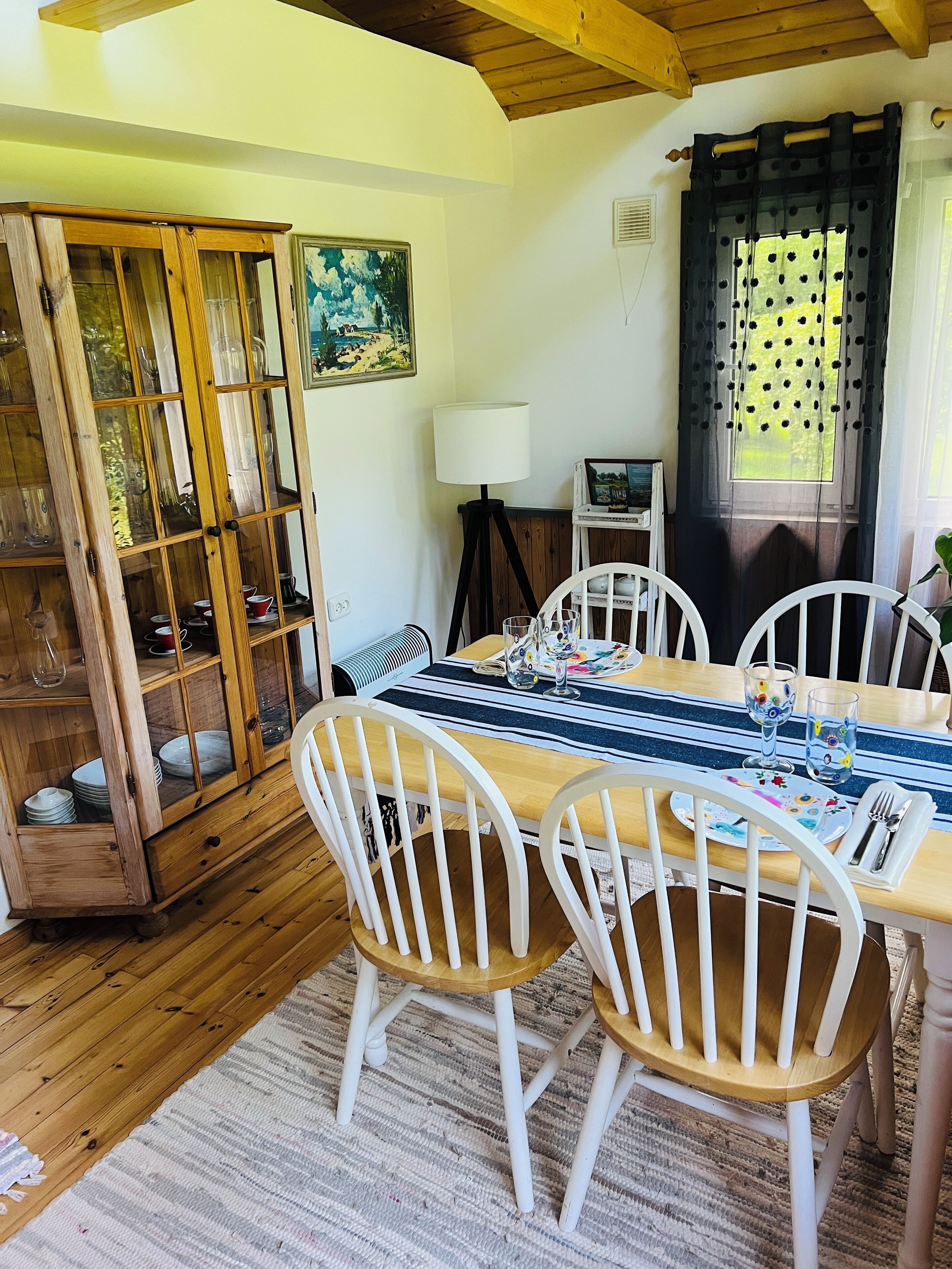 Zona pranzo con tavolo in legno, sedie bianche, credenza in vetro con stoviglie, una lampada e una finestra con tende.