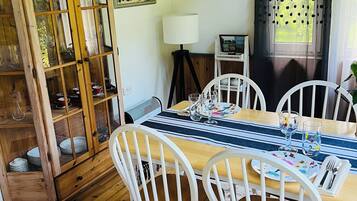 Zona pranzo con tavolo in legno, sedie bianche, credenza in vetro con stoviglie, una lampada e una finestra con tende.