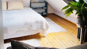 A bedroom with a sloped ceiling, a bed with white bedding, a bedside table with a lamp, a small plant, and a view through a window.