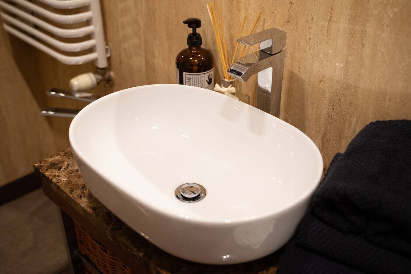 A white oval sink with a modern faucet, a soap dispenser, and reed diffusers on a wooden vanity.