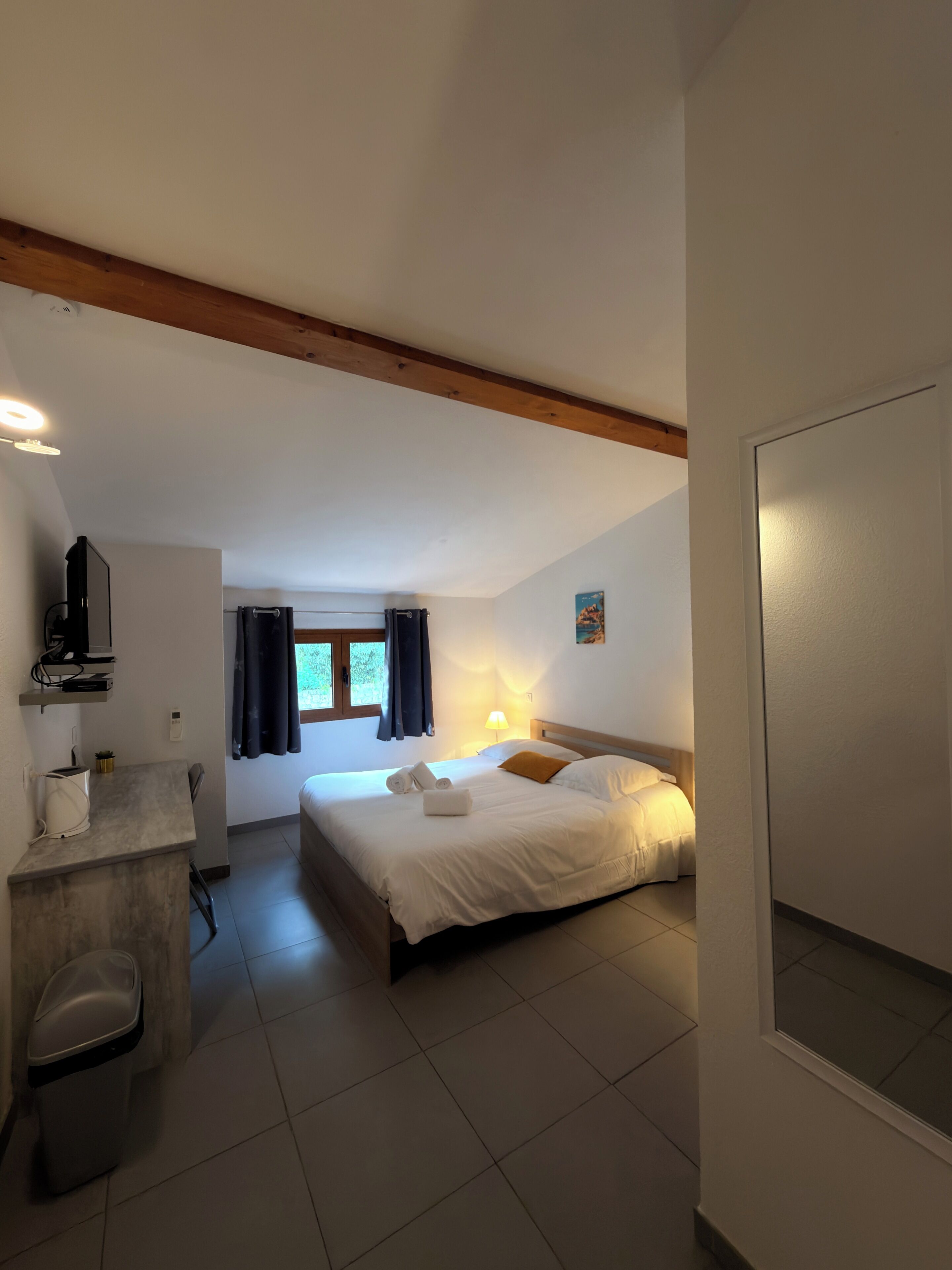 A hotel room with a bed, bedside table, lamp, and a view of the outdoors.