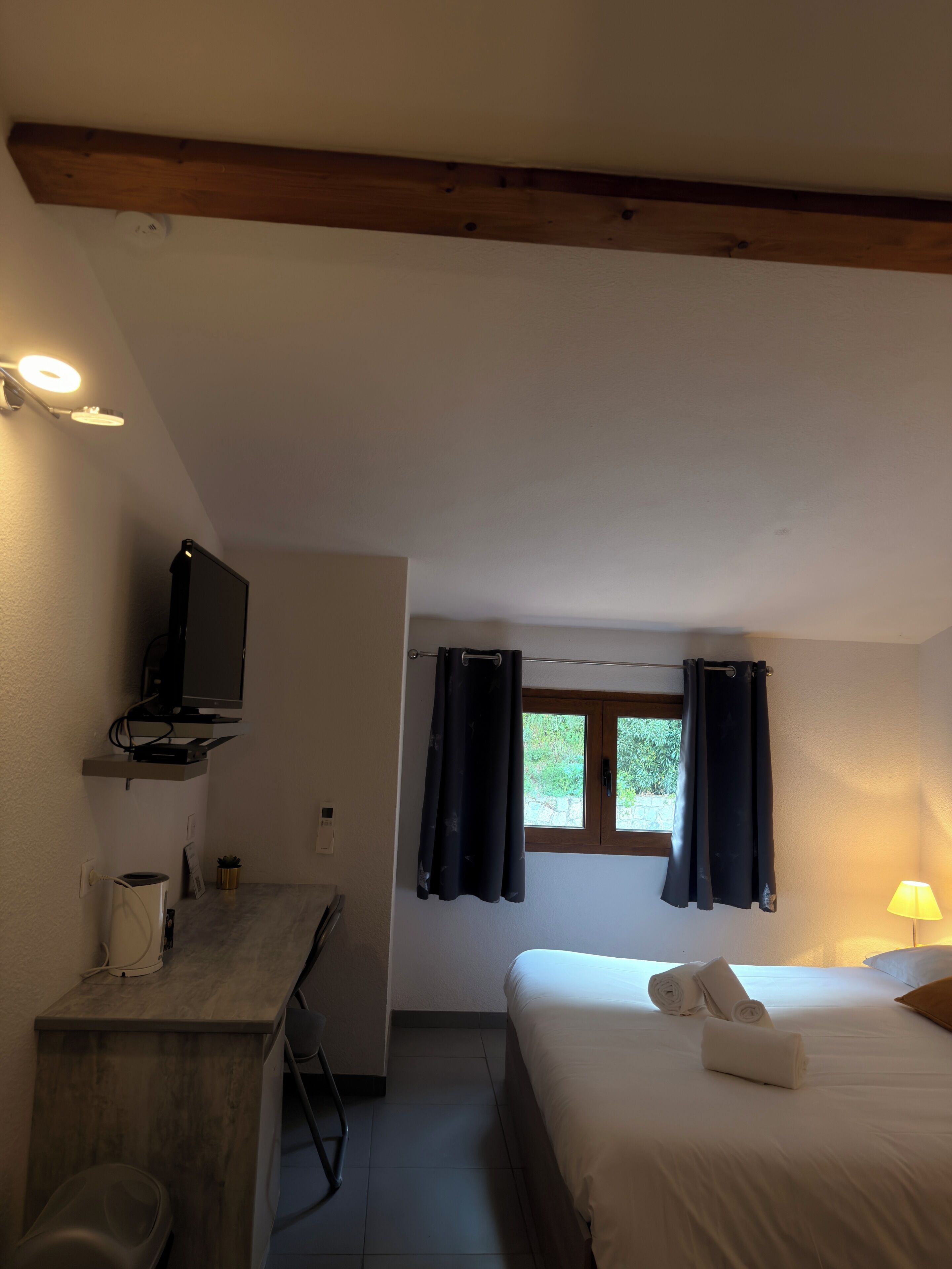 Una habitación con cama, un escritorio con computadora, una ventana con cortinas y una lámpara.