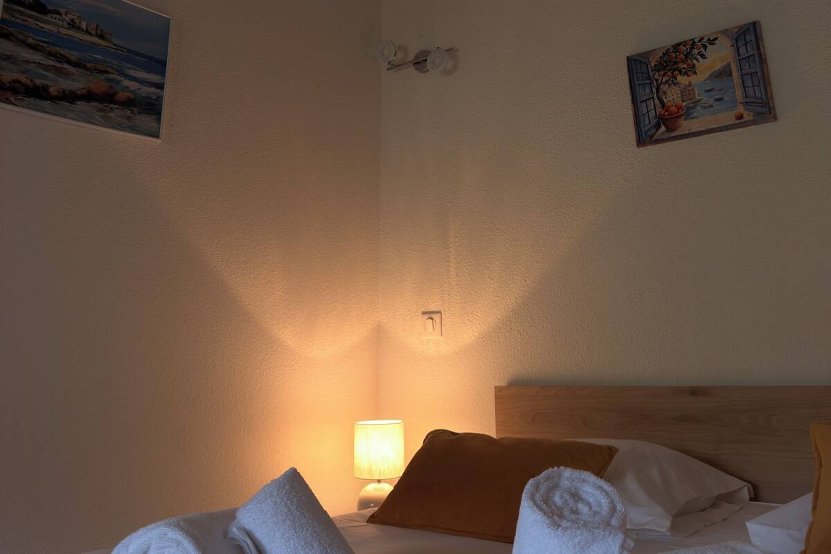 Habitación de hotel con cama, dos toallas, mesita de noche con lámpara y dos cuadros en las paredes.