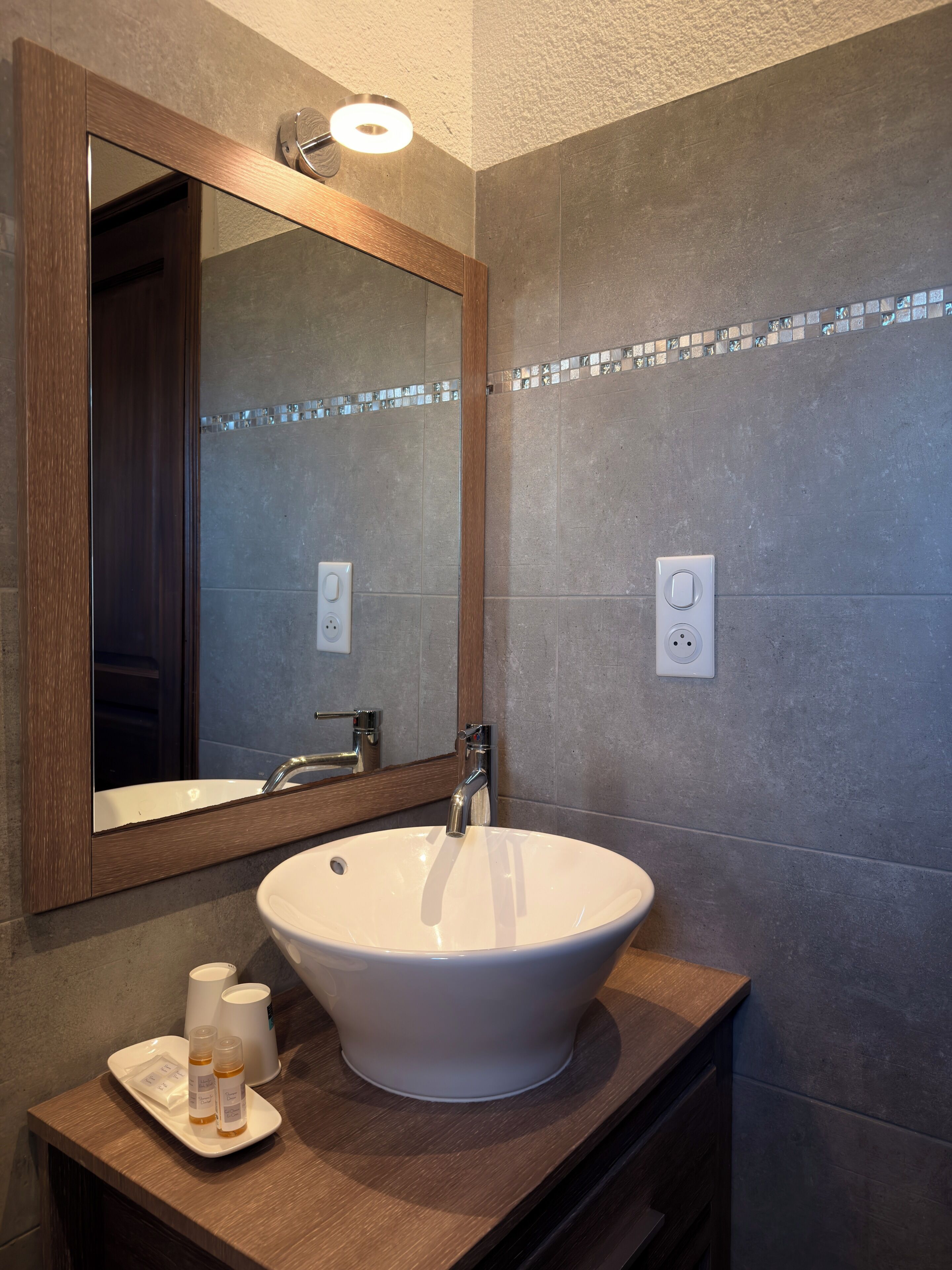 Ein Badezimmer mit einem großen Spiegel, einem Waschtischbecken, einer Holzwaschkommode und einer kleinen Ablage mit Toilettenartikeln.