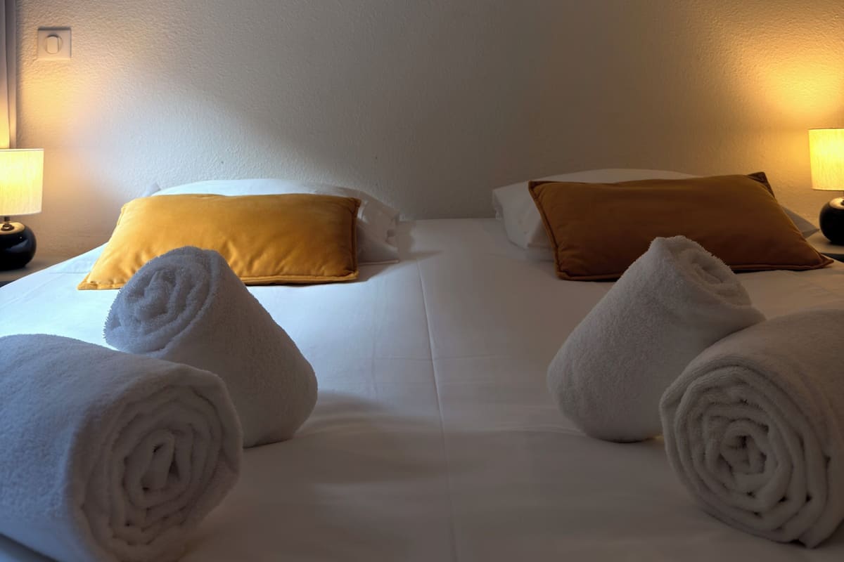 Habitación de hotel con dos camas, ropa de cama blanca, toallas enrolladas, un cuadro en la pared y lámparas de noche.