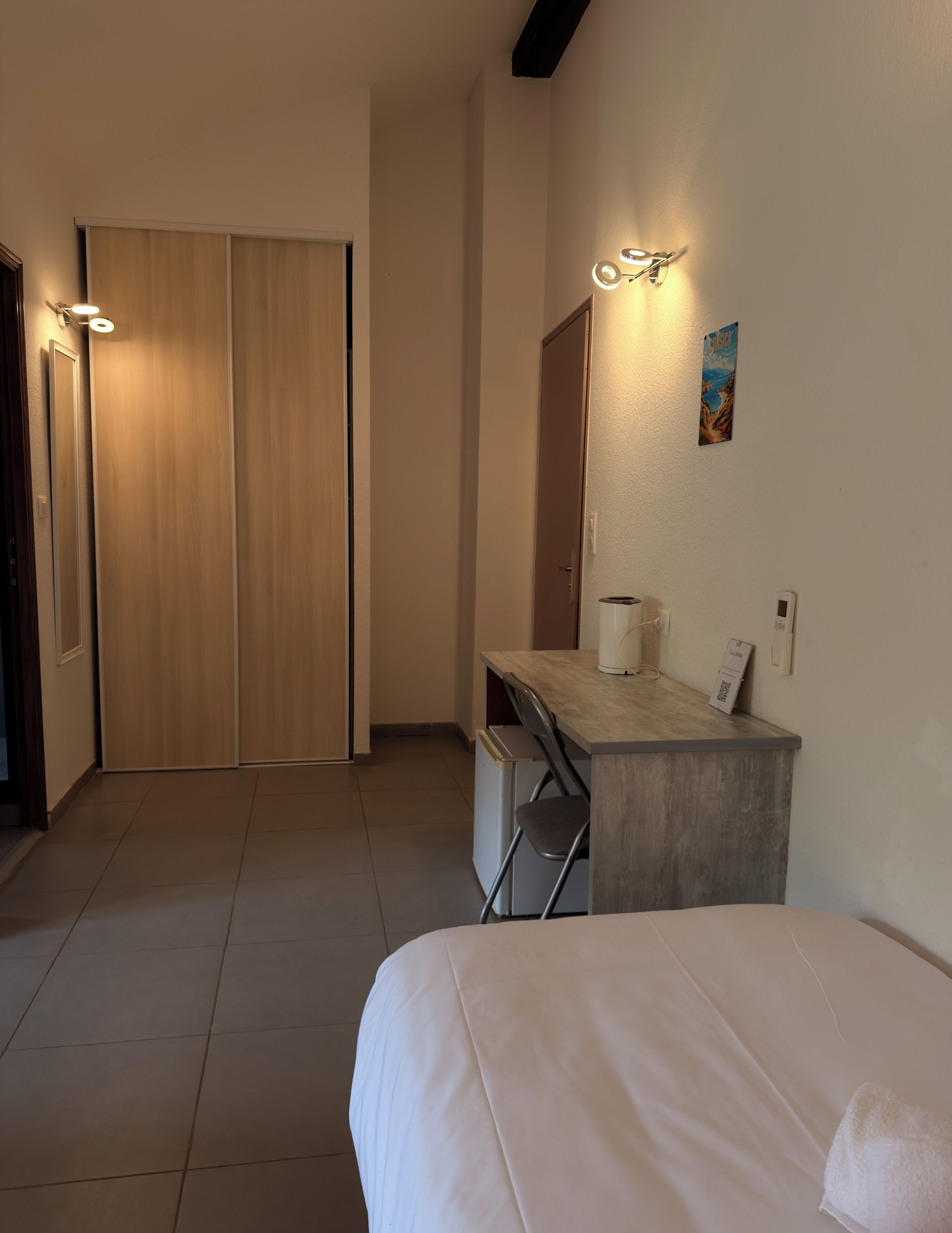 Habitación de hotel con cama, escritorio con silla, armario y lámpara fijada a la pared.