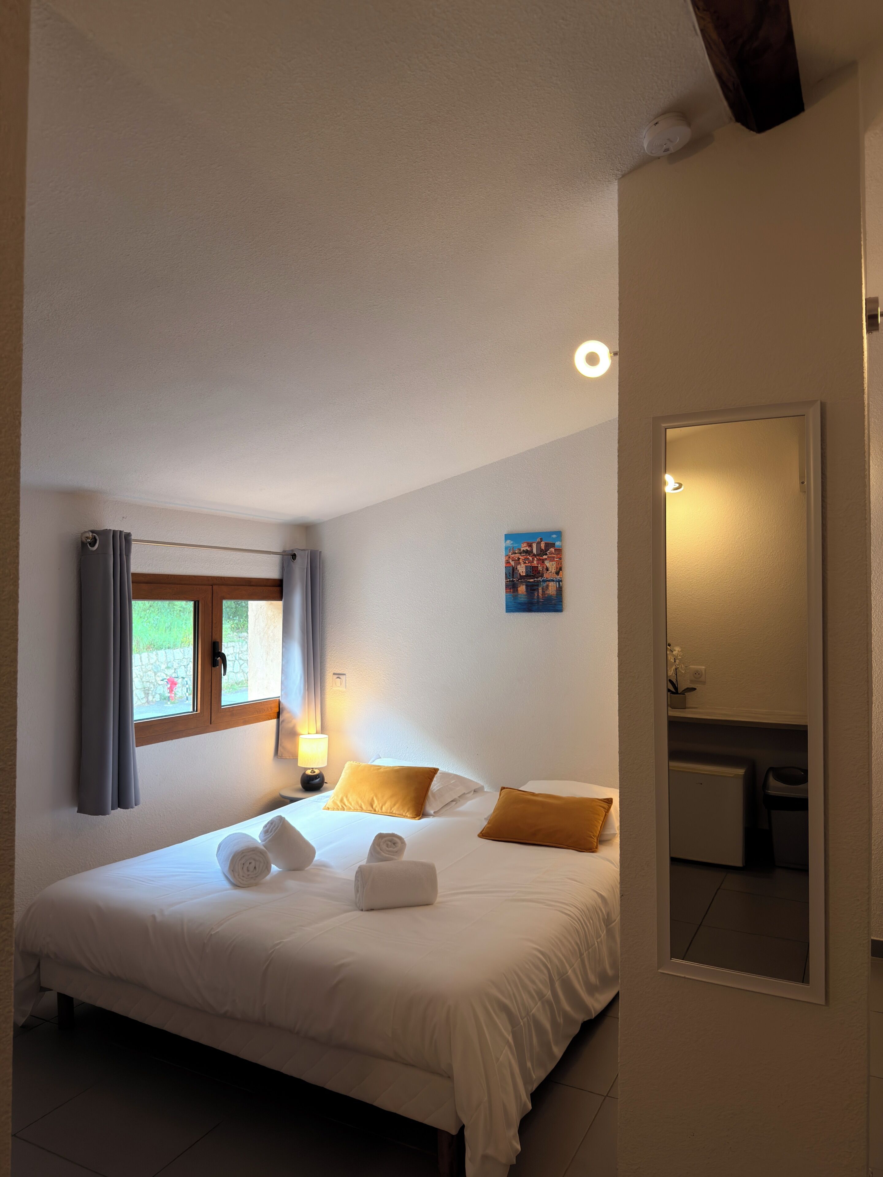 A bedroom with a bed, a window with curtains, a bedside lamp, a small table, and a mirror.