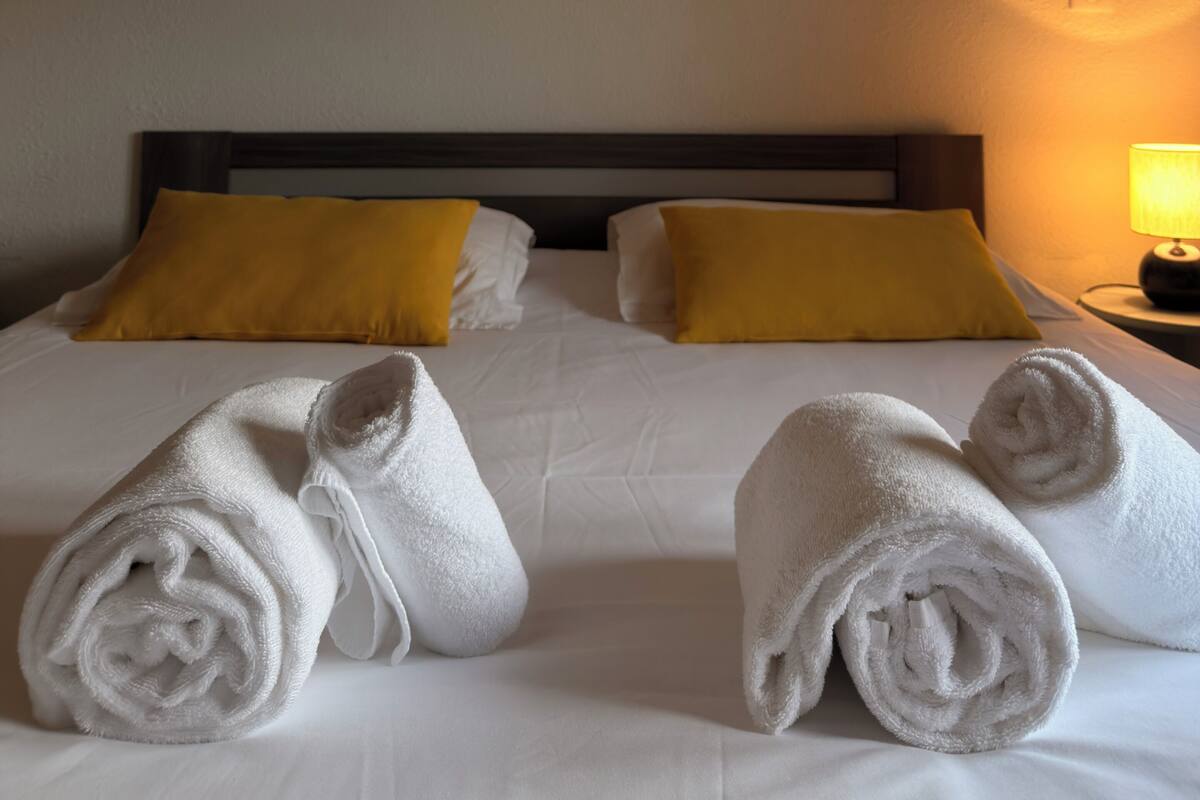 Habitación de hotel con cama, dos almohadas amarillas, mesita de noche con lámpara y un cuadro en la pared.