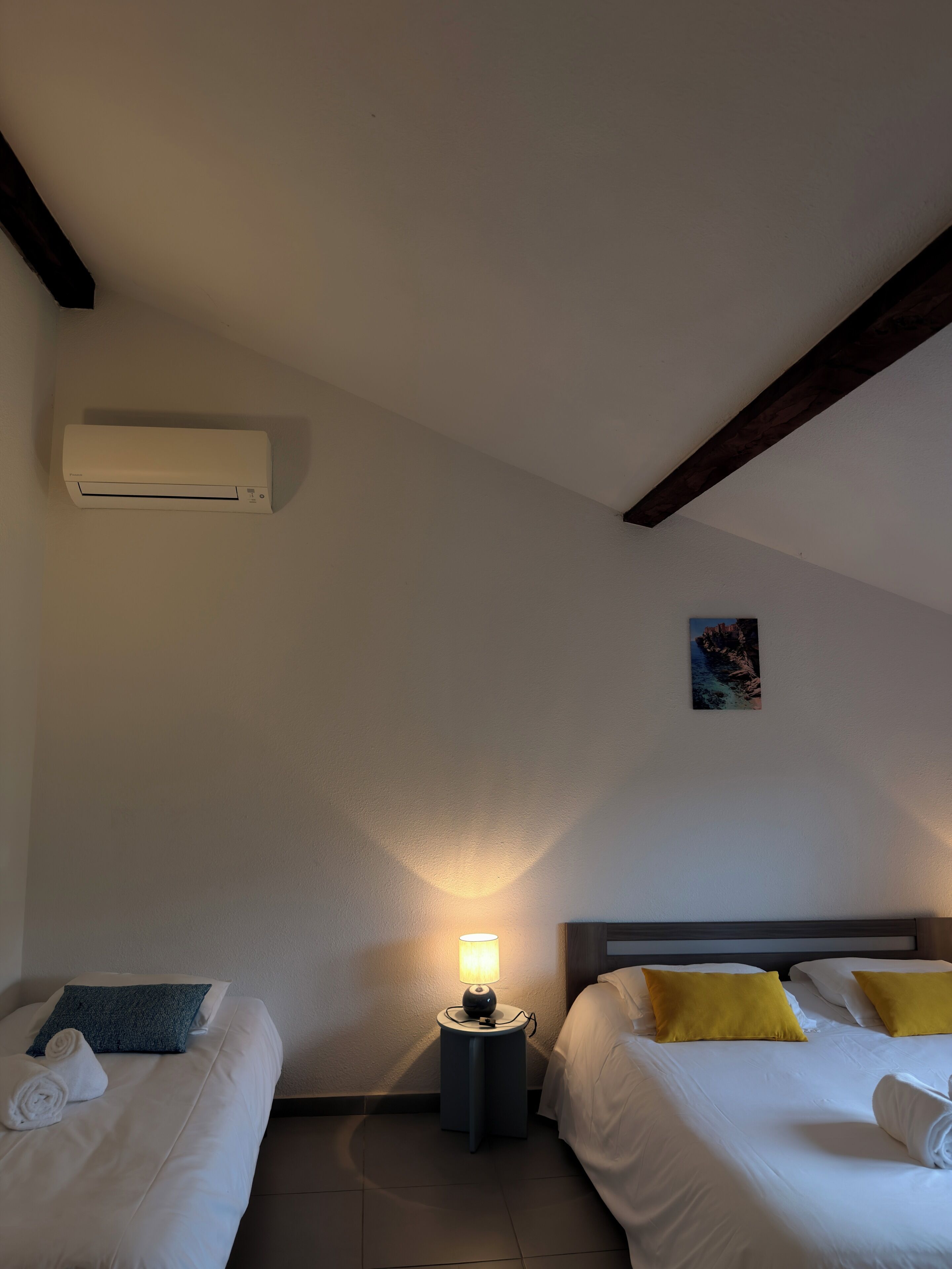 Hotellihuone, jossa on kaksi sänkyä, yöpöytä lampun kanssa, ilmastointilaite ja pieni taulu seinällä.