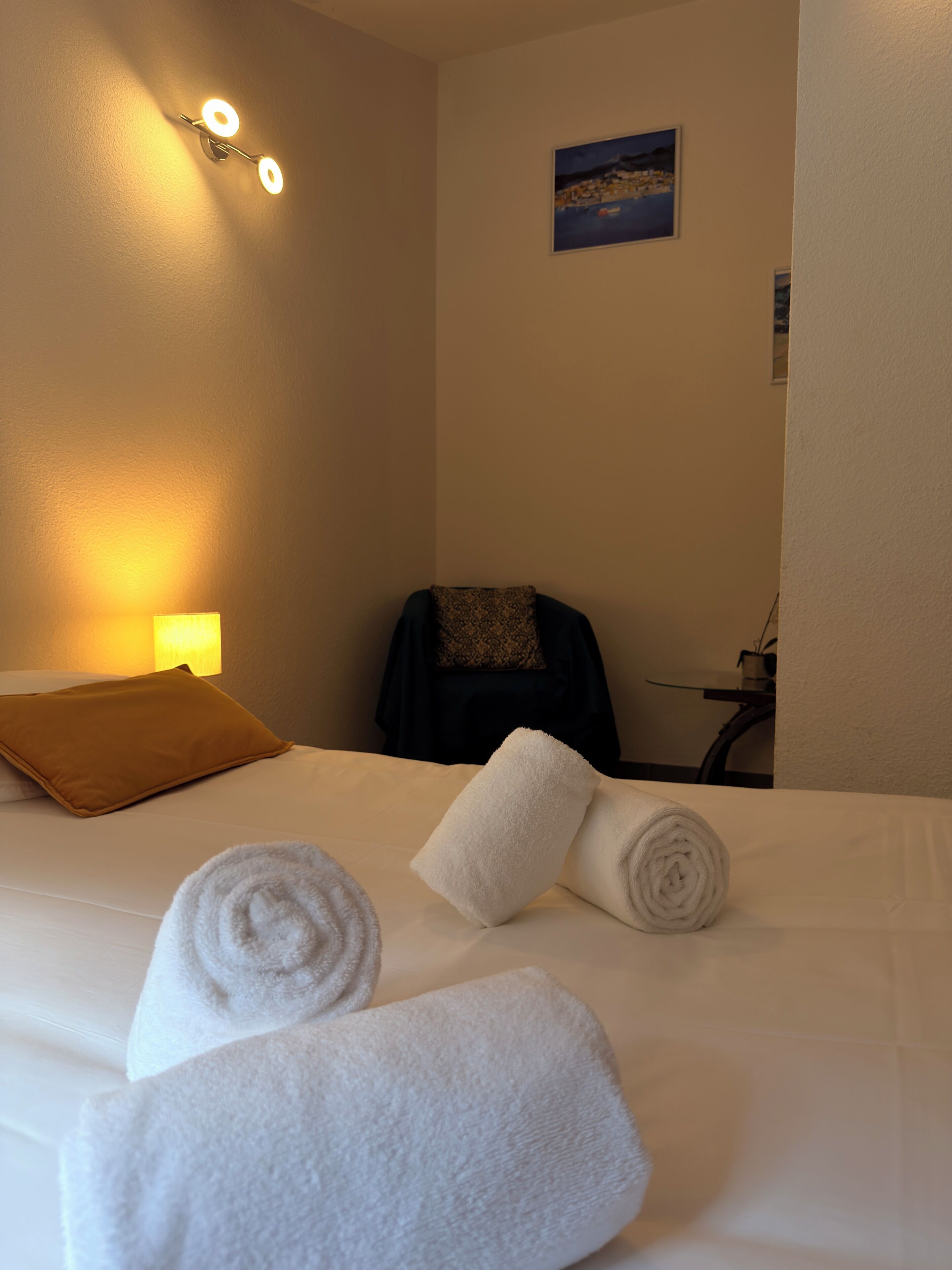 Habitación de hotel con cama, dos toallas enrolladas, una silla, un escritorio, una lámpara y un cuadro en la pared.