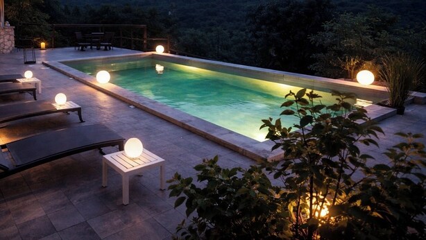A rectangular pool with a stone border, surrounded by lounge chairs and small tables with spherical lights, set against a mountainous backdrop. - Casa Stella  (Olmeto)