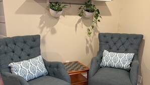 Een kamer met twee grijze fauteuils met gestreepte kussens, een klein houten bijzettafeltje en een passend poefje. Boven het tafeltje staan twee potplanten op witte planken.