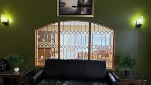 Une pièce avec un canapé en cuir noir, une table en bois ornée d’un pot de plantes, une grille décorative à la fenêtre et un tableau encadré accroché au mur.