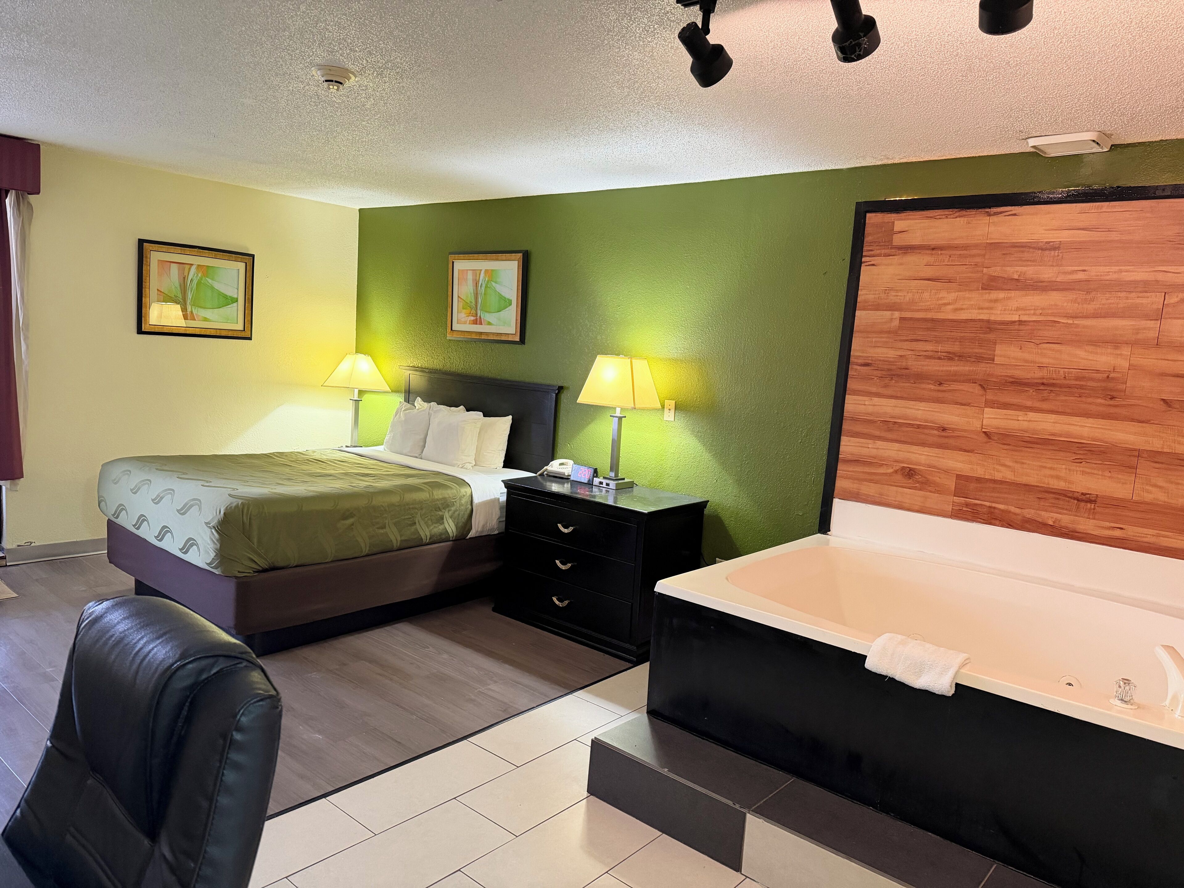 A hotel room with a bed, bedside lamps, a wooden headboard, a black nightstand, a black chair, and a large bathtub.