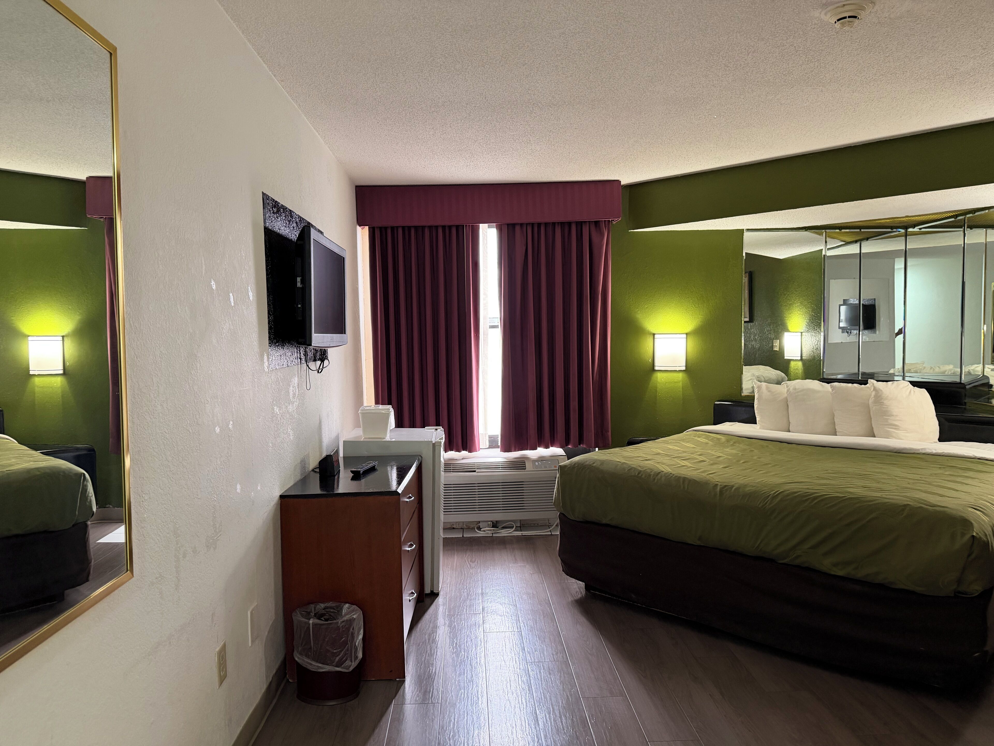 A hotel room with a bed, a desk with a telephone, a trash bin, a television mounted on the wall, and a window with curtains.