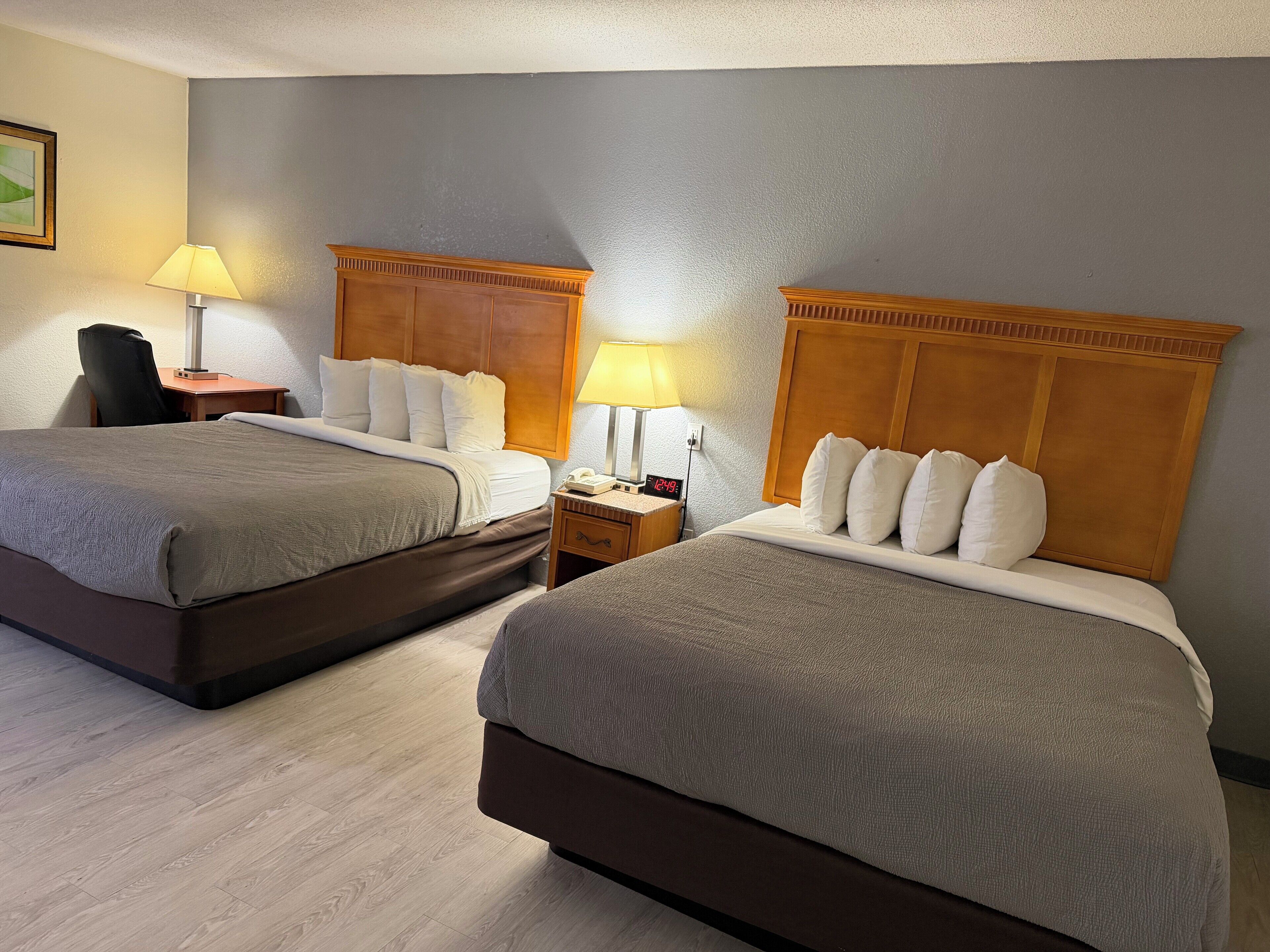 A hotel room with two beds, a nightstand with a lamp, a chair, and a clock on the wall.