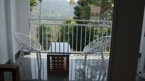 Un balcón con dos sillas blancas y una pequeña mesa, con vistas a un paisaje verde y exuberante con palmeras y montañas.