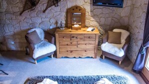A stone-walled room with two fur-covered chairs, a wooden dresser with drawers, a mounted television, and a small mirror.