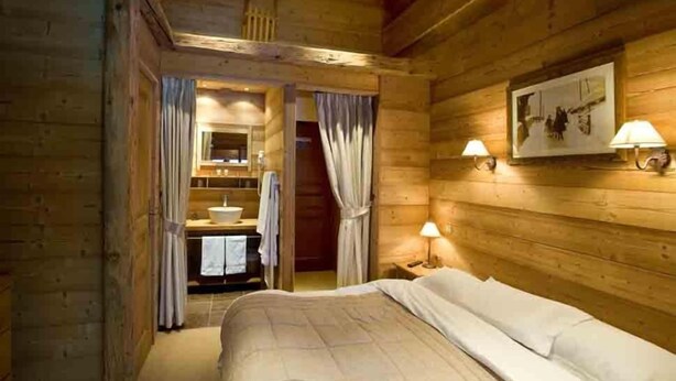 A wooden cabin room with a bed, a bathroom with a sink, and two wall-mounted lamps.