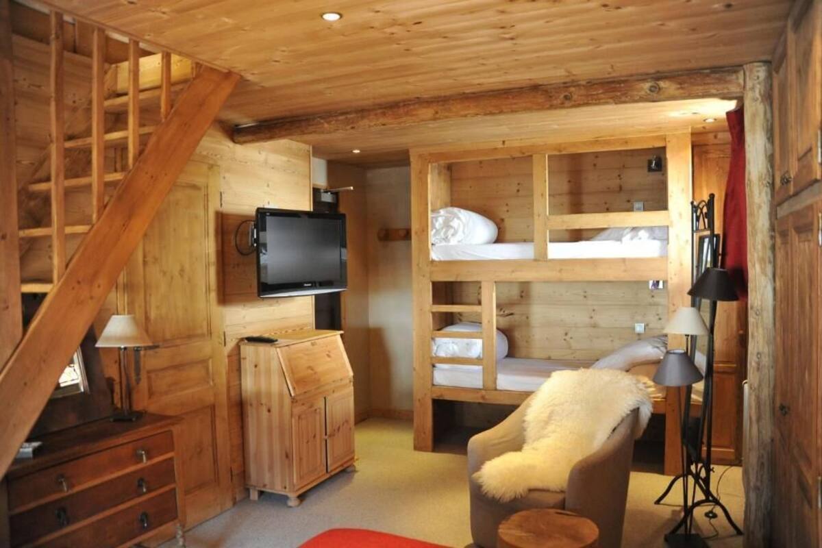 Un accogliente interno di una baita con una scala in legno, una televisione, letti a castello, una poltrona con una coperta in pelliccia e una lampada.