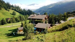 A rural house with a large roof and a smaller attached structure, surrounded by lush green fields and trees.
