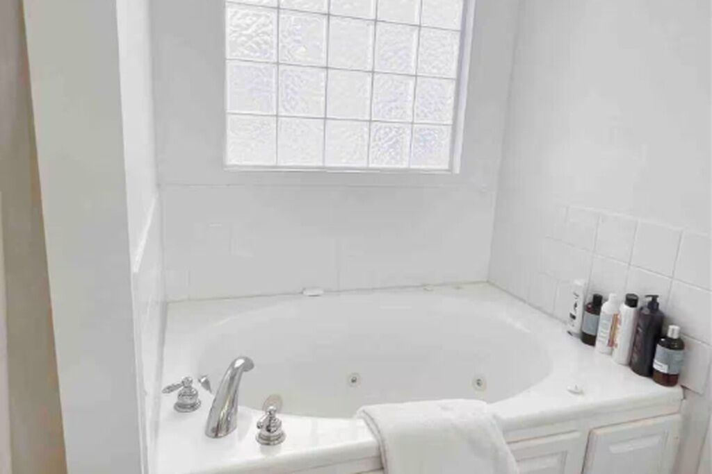 Een witte badkamer met een vrijstaand bad, een raam met gezandstraald glas en toiletartikelen aan de wand.