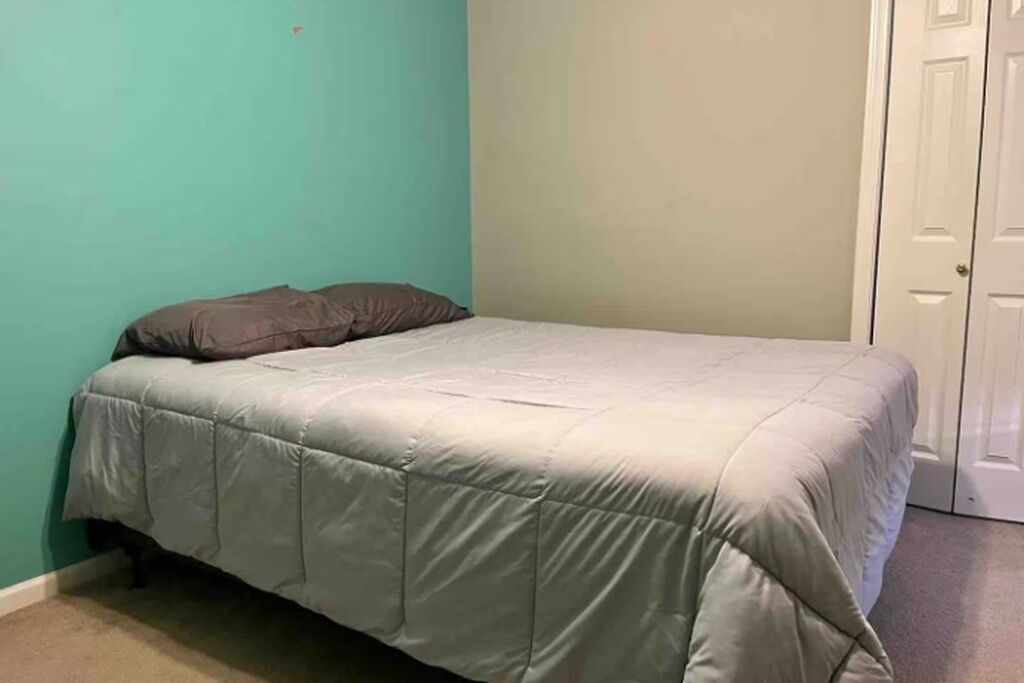 Un lit simple avec une couvre-lit grise dans une pièce aux murs de couleur turquoise et à la porte blanche.
