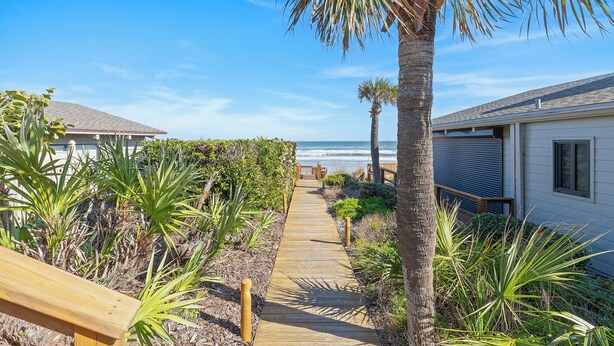 Un sentier en bois mène à une maison de plage offrant une vue sur l’océan.