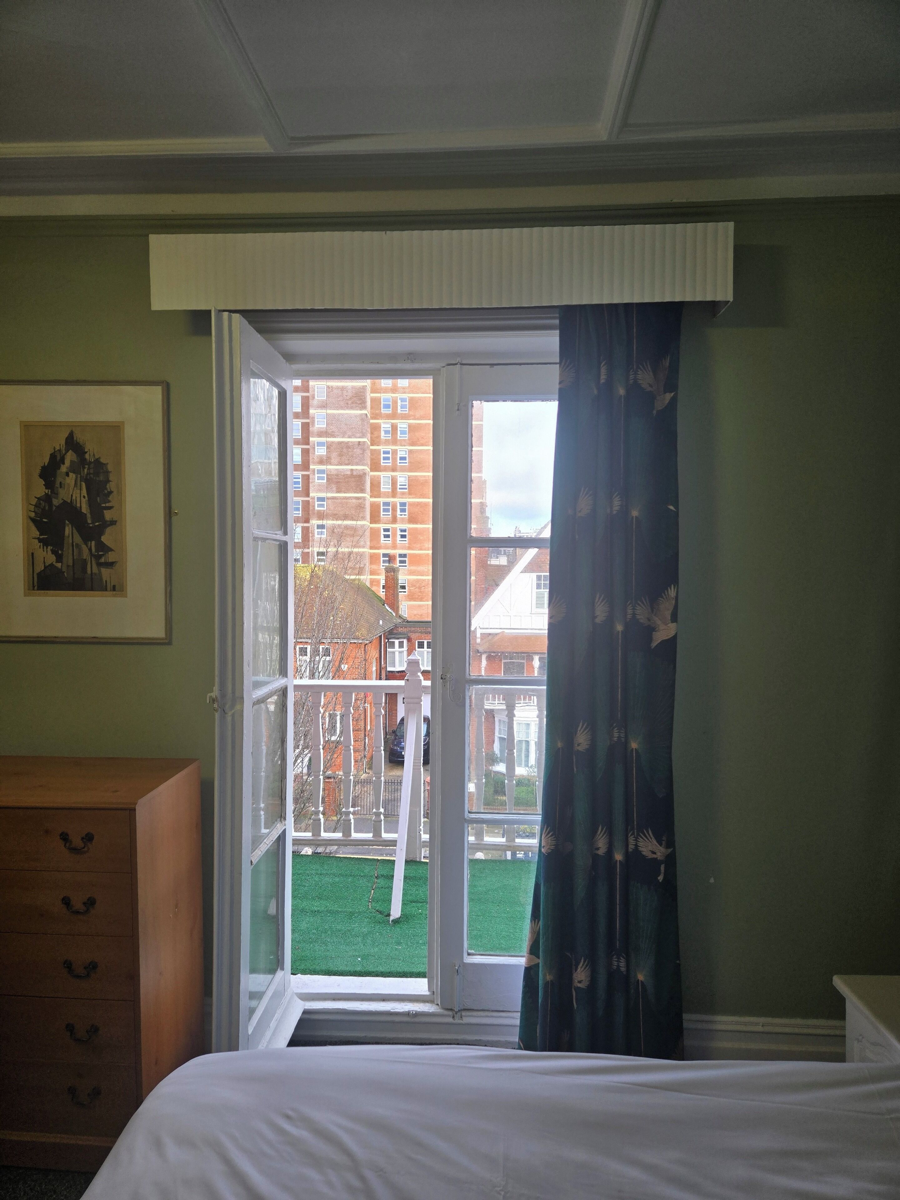 A hotel room with a bed, a wooden dresser, a framed picture on the wall, and a view of a balcony with a green lawn and a brick building in the background.