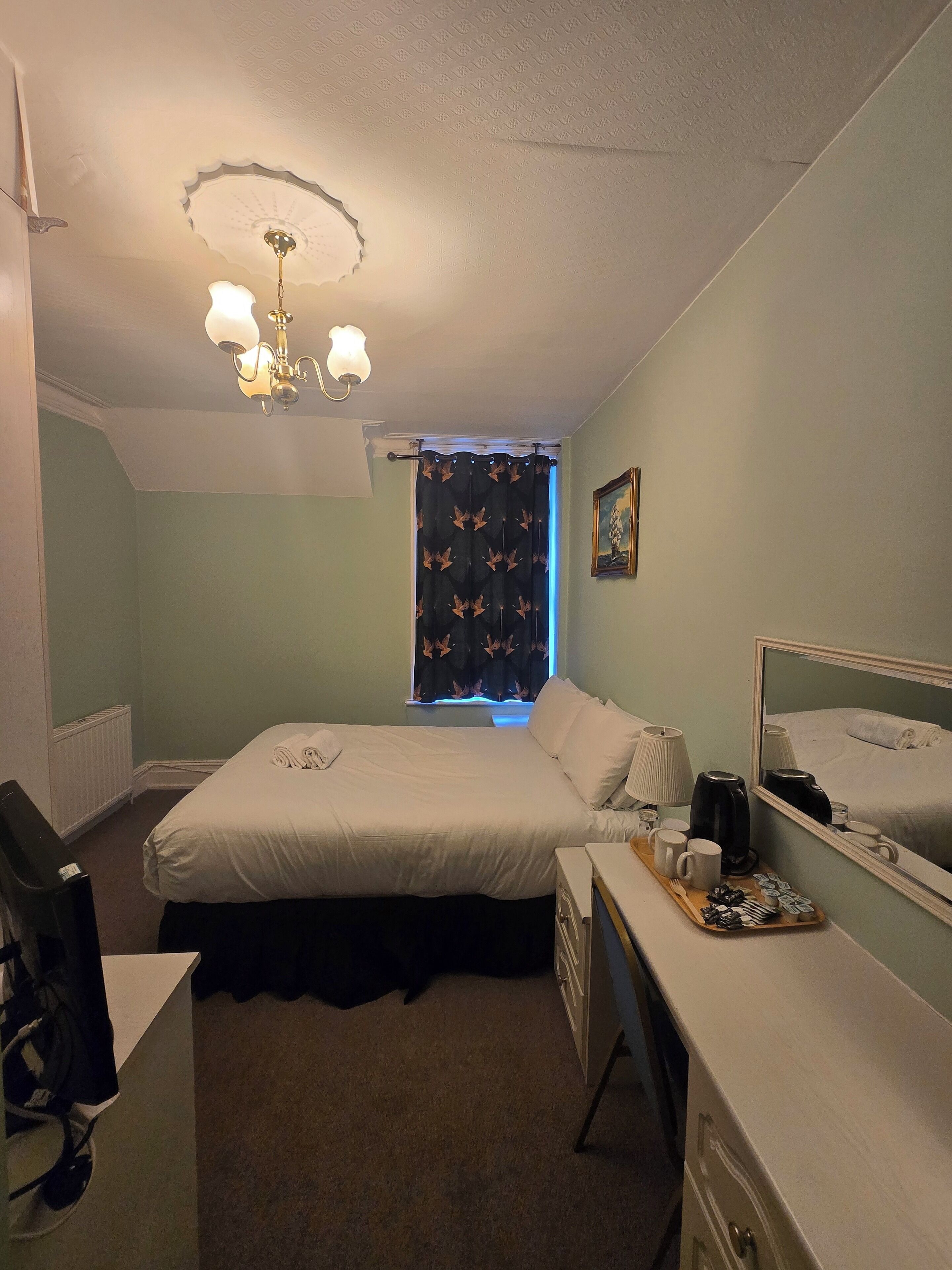 A hotel room with a bed, a desk, a chair, a dresser, a mirror, and a window with a curtain.