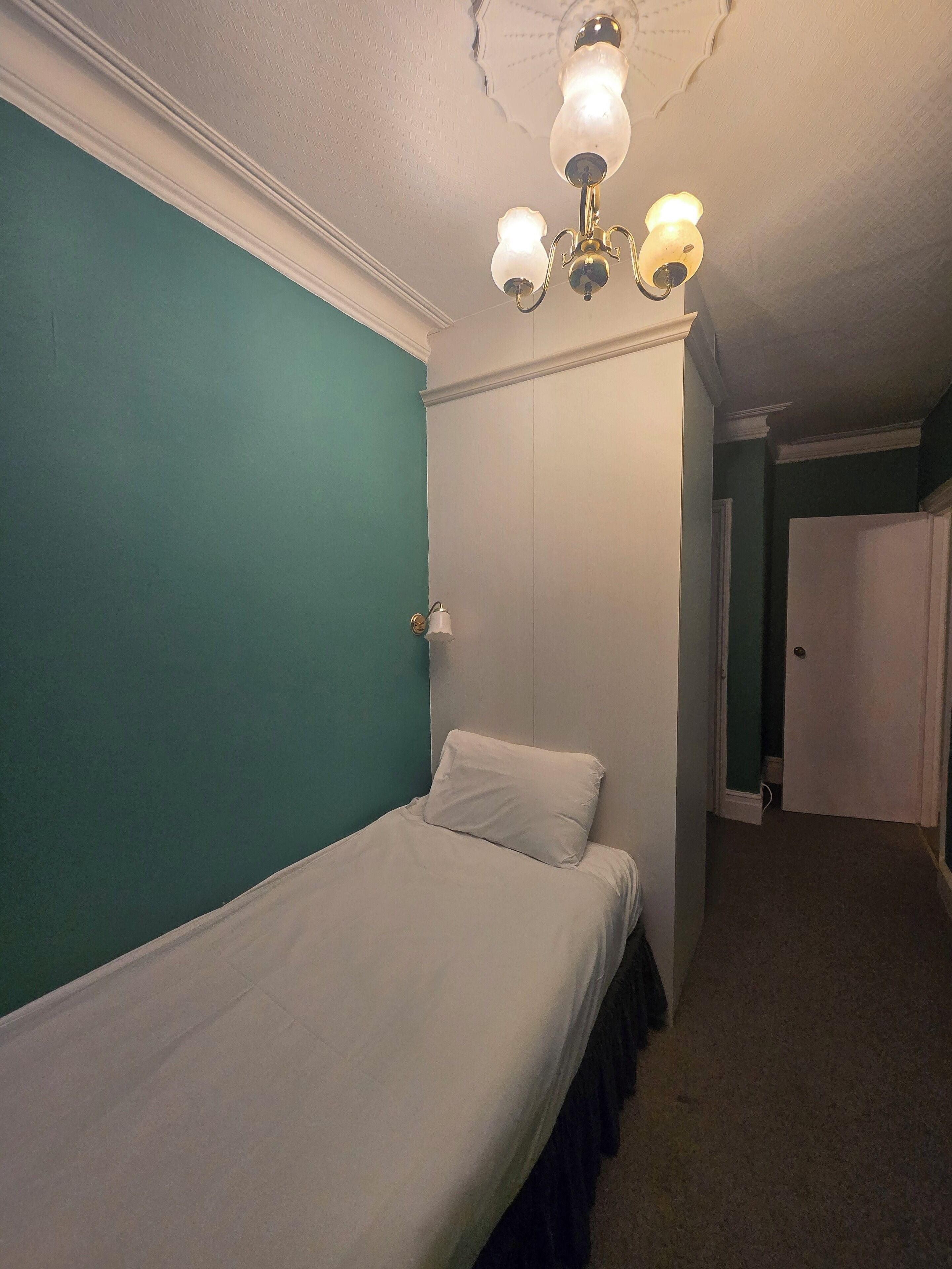 A small, narrow room with teal walls, a single bed with white bedding, and a ceiling light fixture.