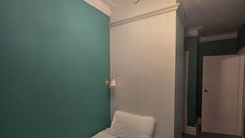 A small, narrow room with teal walls, a single bed with white bedding, and a ceiling light fixture.