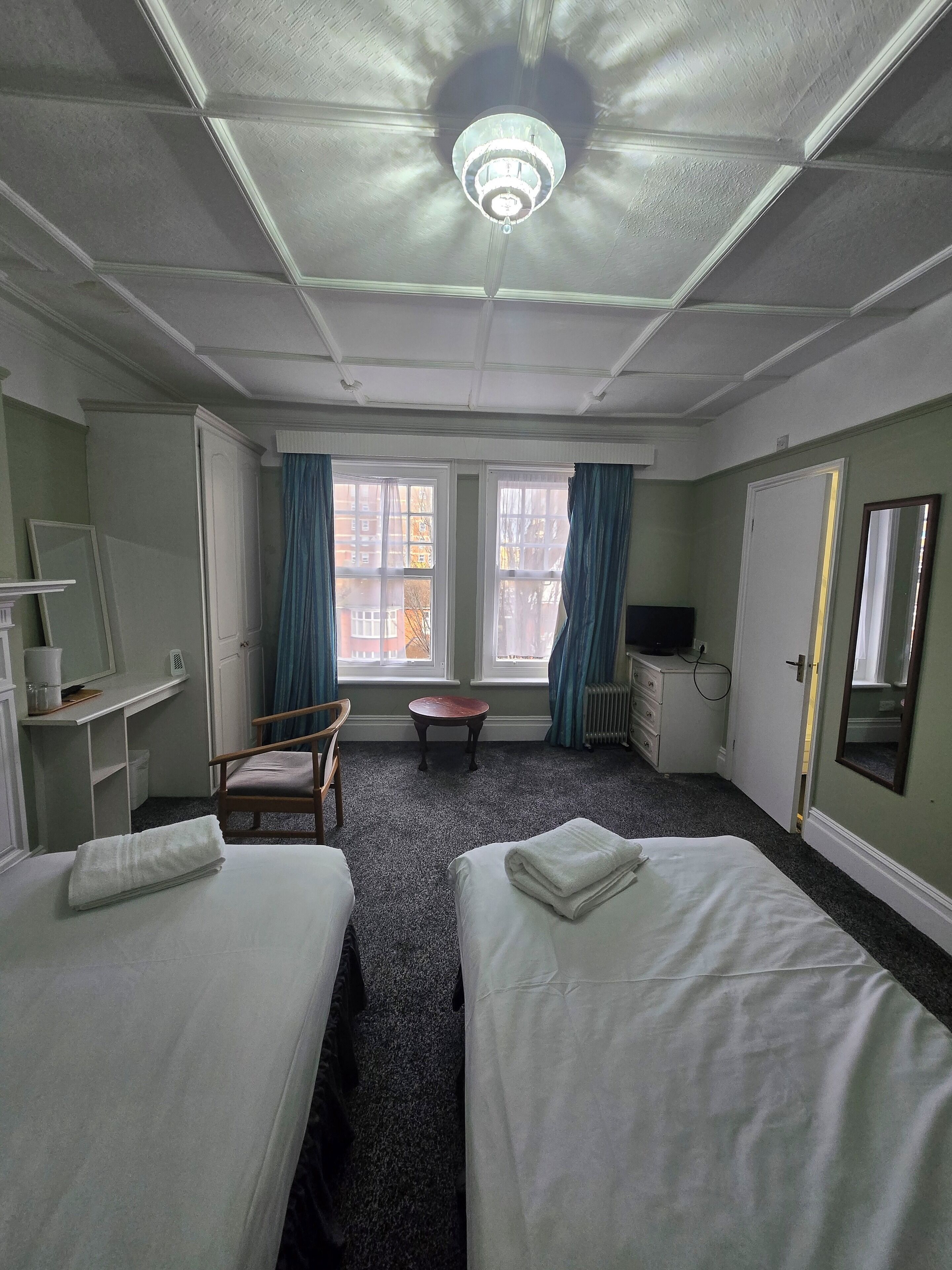 Et hotelværelse med to senge, et skrivebord, en stol, et lille bord og en loftlampe.