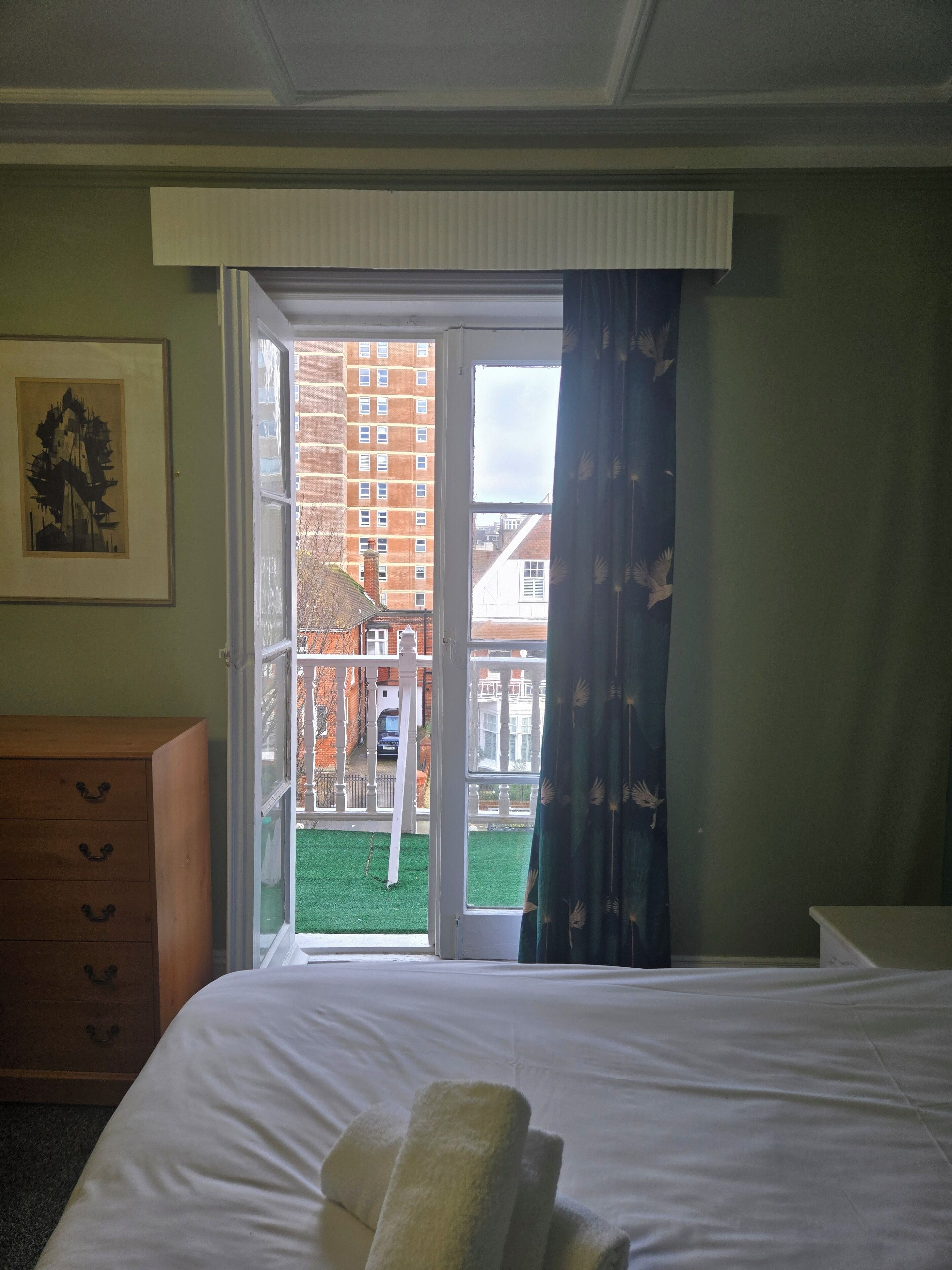 A hotel room with a bed, a wooden dresser, a framed picture on the wall, and a view of a building through an open door.