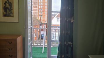 A hotel room with a bed, a wooden dresser, a framed picture on the wall, and a view of a building through an open door.