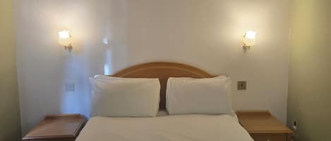 A bedroom with a large bed, two bedside tables, wooden headboard, and wall-mounted light fixtures.