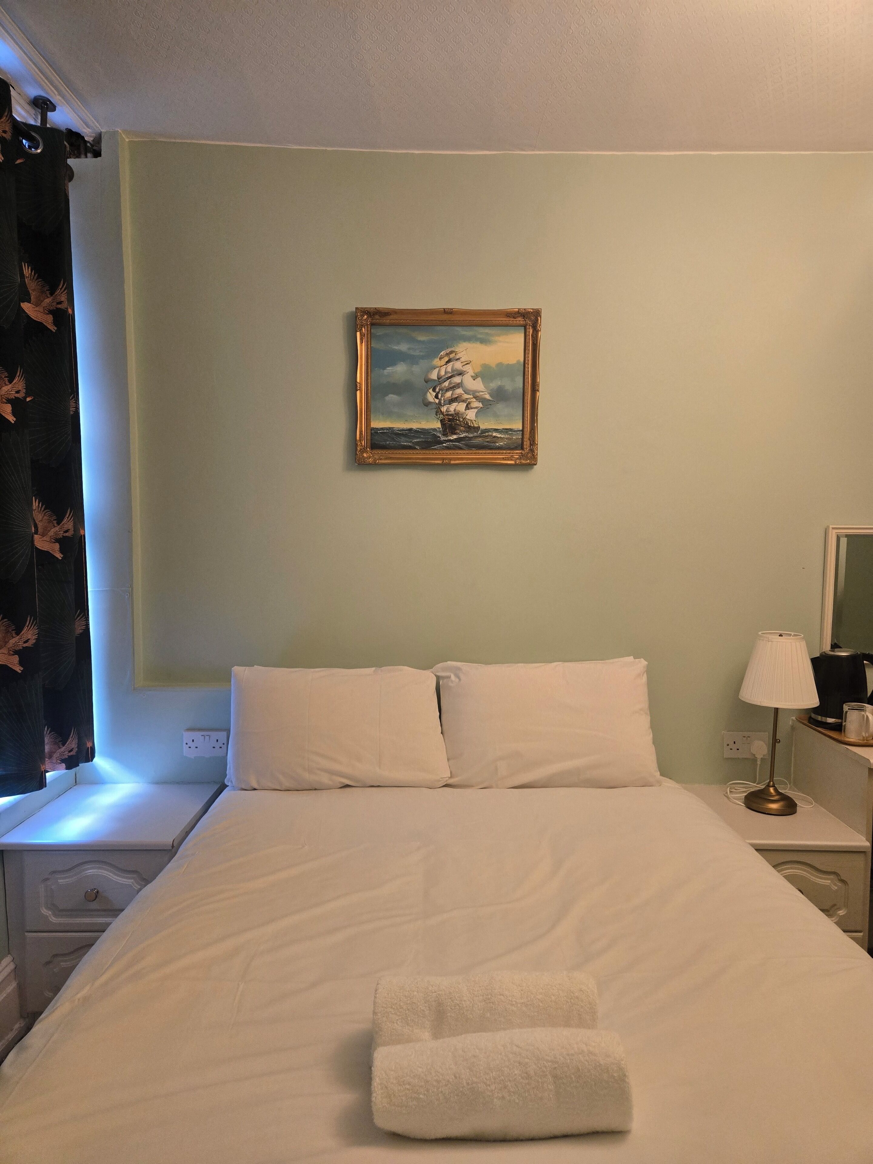 Una habitación de hotel con cama, almohadas, un cuadro de un barco en la pared, una mesita de noche con lámpara y una ventana con cortinas.