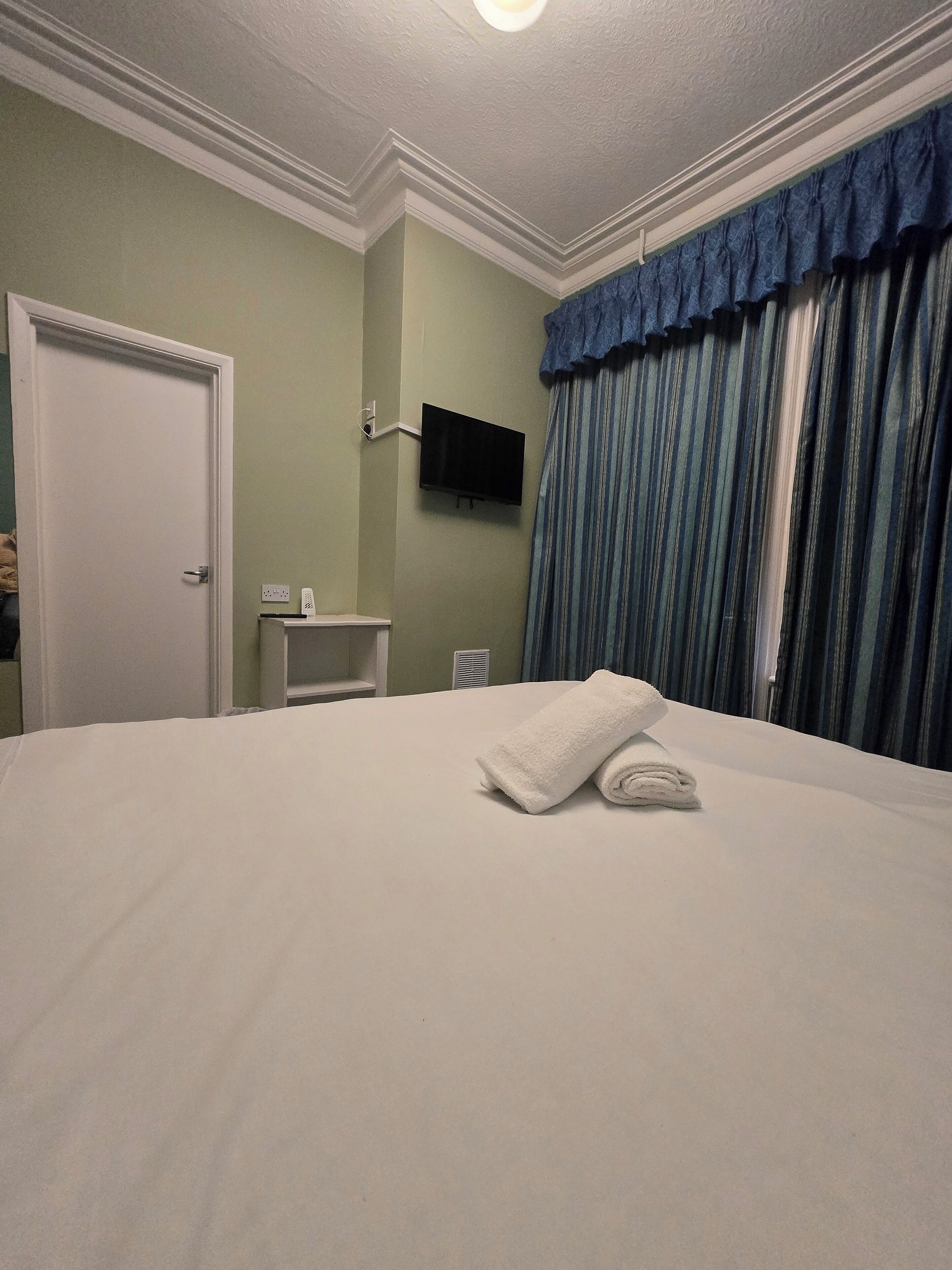 Quarto de hotel com cama, televisão de tela plana na parede, mesa de cabeceira, porta e janela com cortinas.