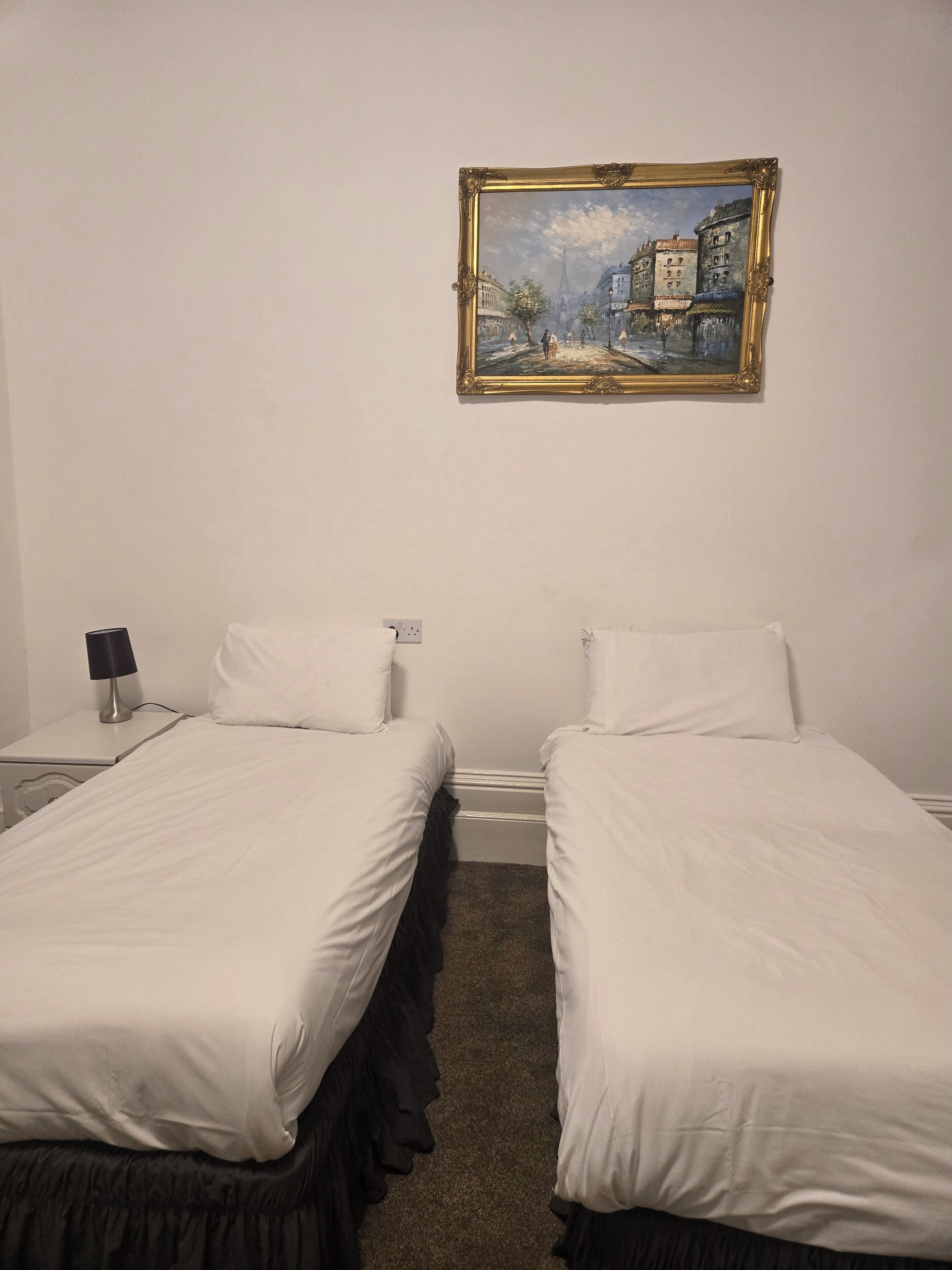 A hotel room with two single beds, a nightstand with a lamp, and a painting on the wall.