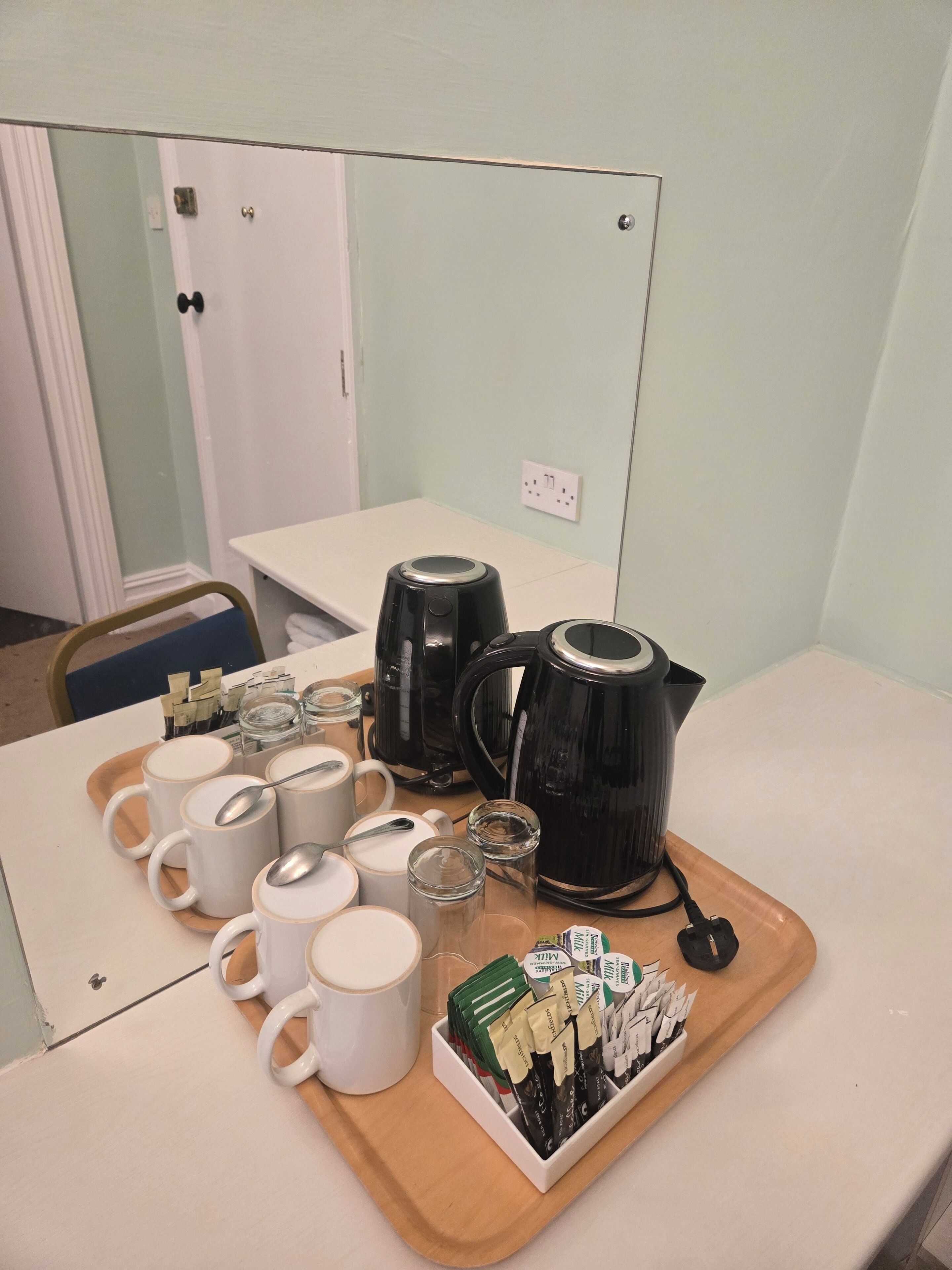 Una bandeja de desayuno con cafeteras, tazas y sobres de café sobre una mesa en una habitación con una puerta y un interruptor de luz en la pared.