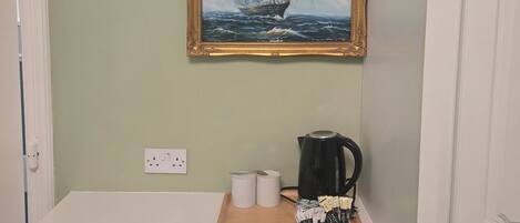 Una habitación con escritorio, un cuadro enmarcado de un barco en la pared, un hervidor y algunas toallas enrolladas.