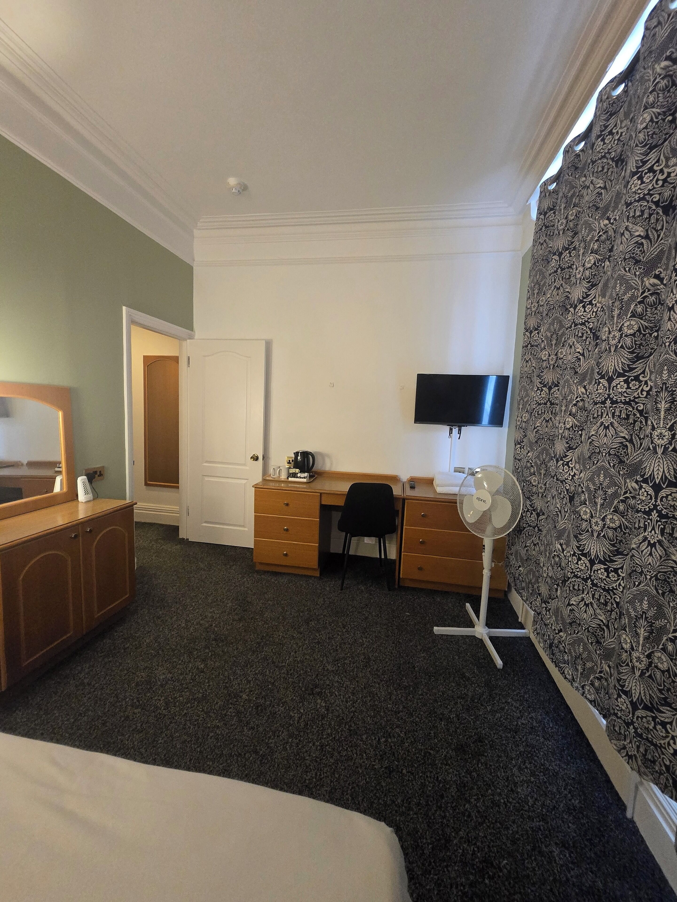 A hotel room with a bed, a desk with a chair, a television, a desk fan, and a wardrobe.