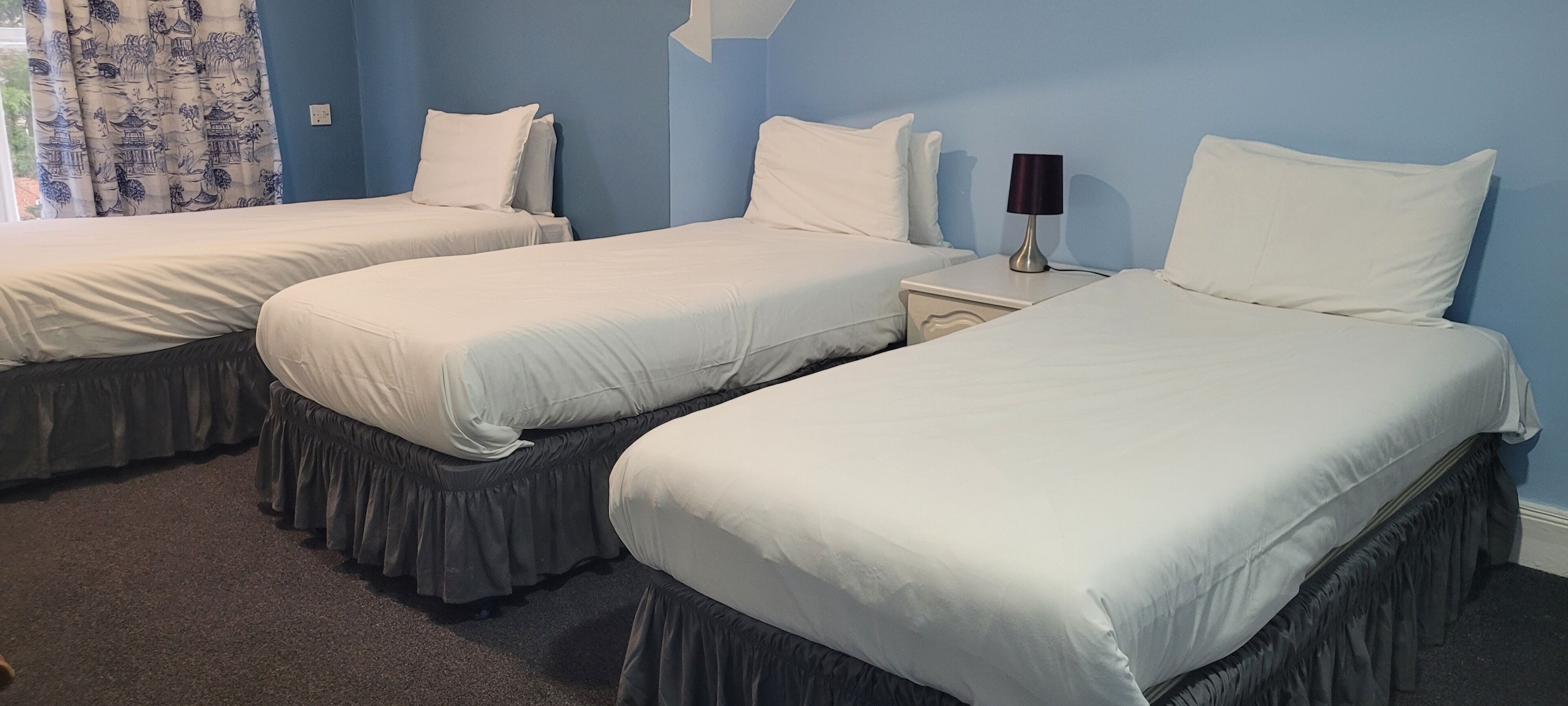 Quarto de hotel com três camas de solteiro, cada uma com um cobre-cama branco e almofadas combinando. Há uma mesa de cabeceira com uma luminária entre as camas.