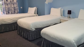 A hotel room with three single beds, each with a white bedspread and matching pillows. There is a nightstand with a lamp between the beds.